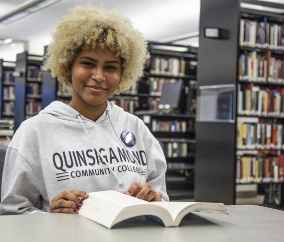 A female student looks up from the book she is reading at a desk in QCC’s Alden Library