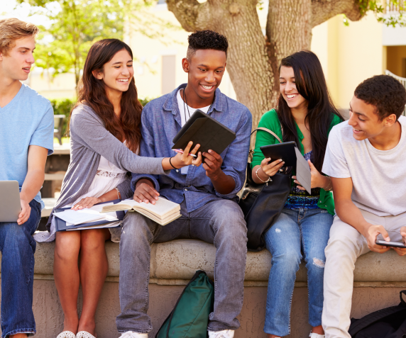 A group of diverse high school students laugh while doing homework out on the lawn