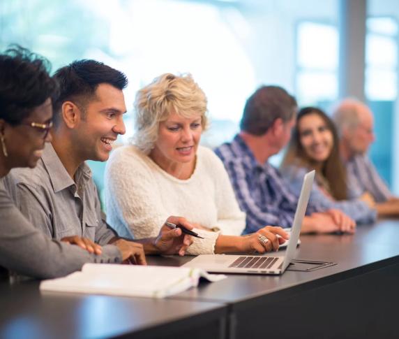 A variety of adult students talk amongst themselves while sitting at a desk in class with laptops