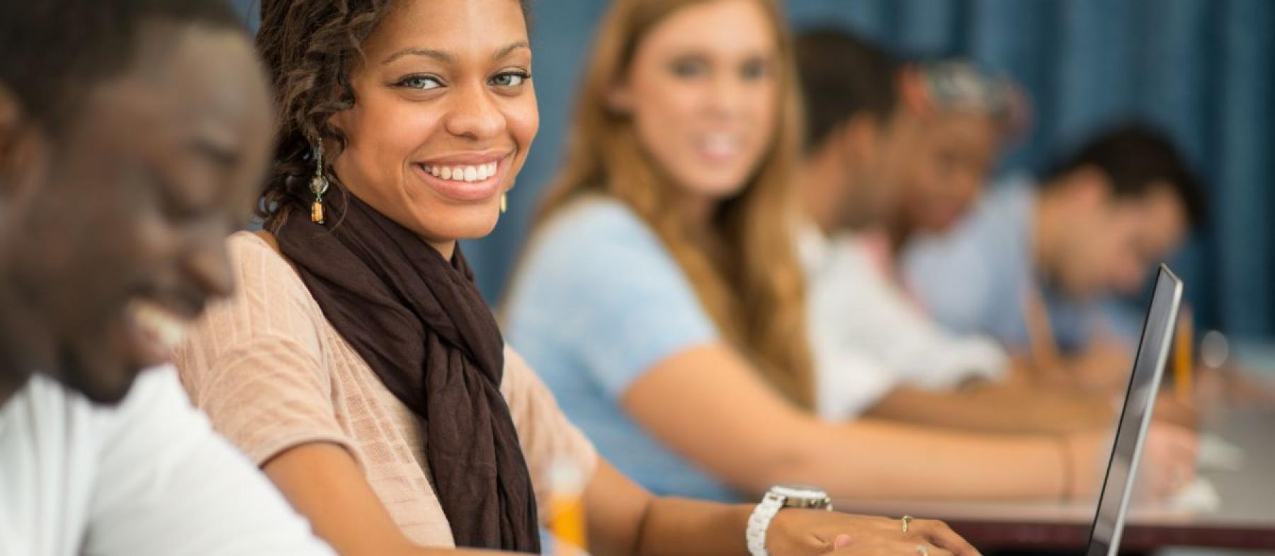 A woman in a classroom with a laptop looks at the camera with a smile