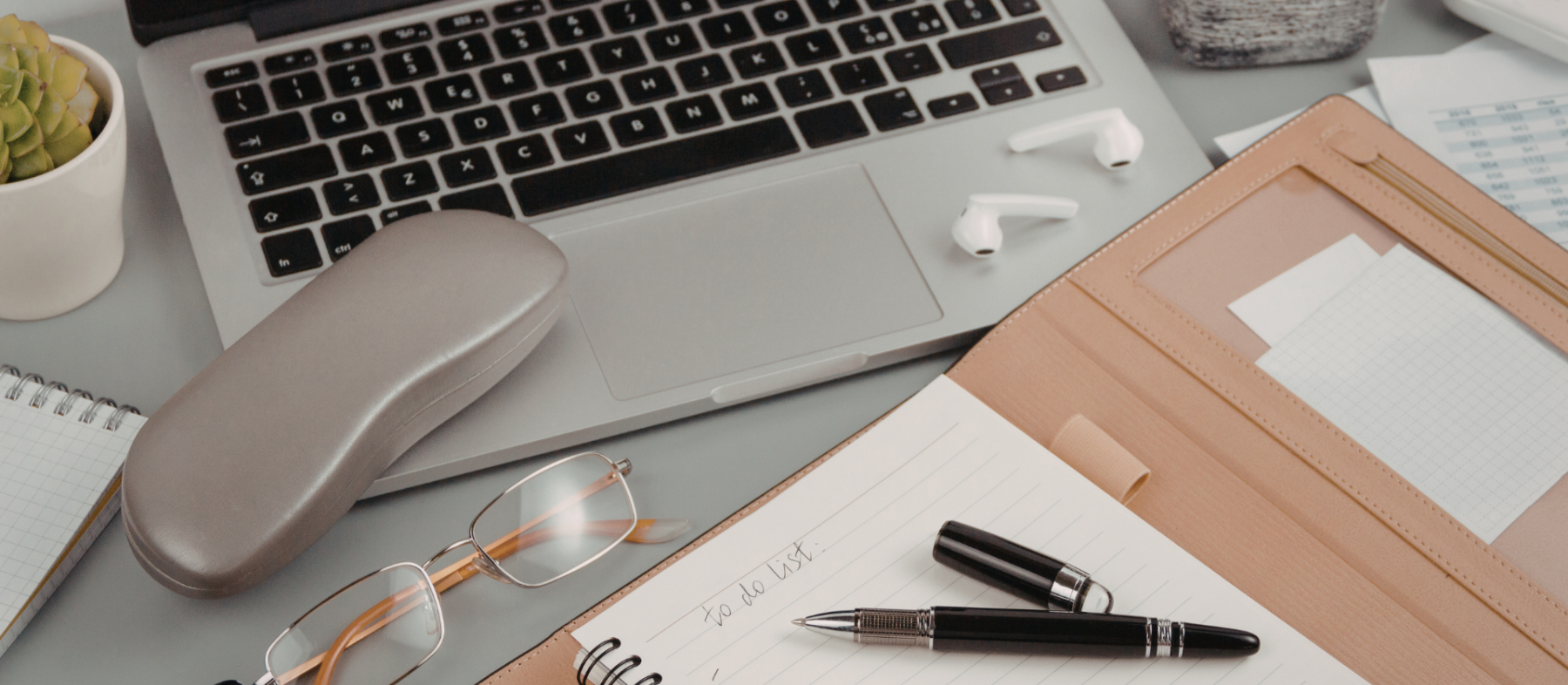 A laptop, glasses case, notebook, and pens lie on a desk.