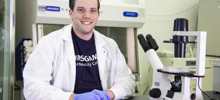 A student wears and lab coat and sits next to a microscope