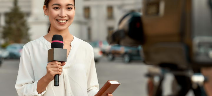 A reporter holds a mic and looks into camera on site